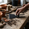 Person using a Carbon Steel Double Headed Garden Hoe-fork on a wooden table with potted plants and gardening tools.