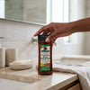 Hand holding Deep Hydrating Licorice Root Glow Body Oil bottle in a modern bathroom setting.