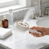 Spray Scalp Massage Comb spraying mist while held above a bathroom countertop with skincare products.