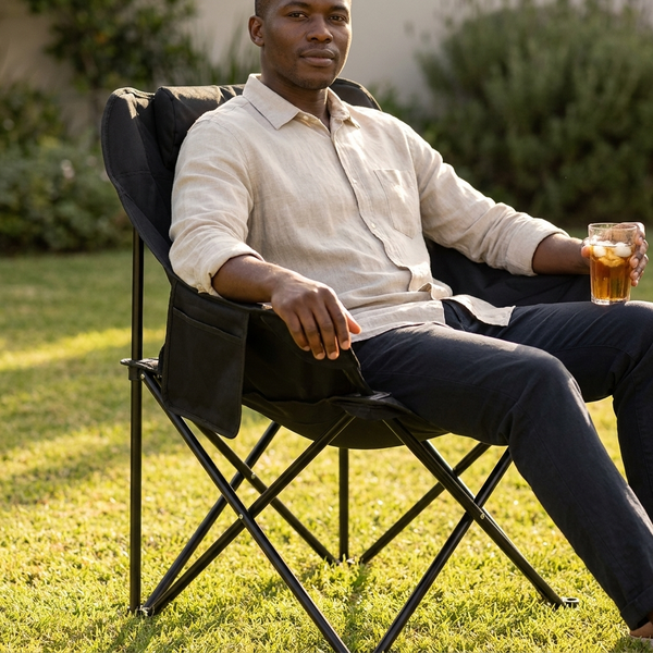 Man relaxing on a Black Folding Moon Chair outdoors, enjoying a drink in a sunny garden.