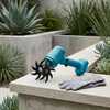 Cordless Handheld Garden Cultivator with rotating tines and gloves on a garden ledge surrounded by plants.