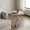 Luna Marble-Look Round Side Table with chrome base, books and a cup on top, next to a light-colored sofa.